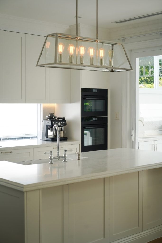 Modern kitchen with a large island, built-in double ovens, espresso machine, and a rectangular pendant light fitting above the island. The space is bright with white cabinetry and natural light from nearby windows.