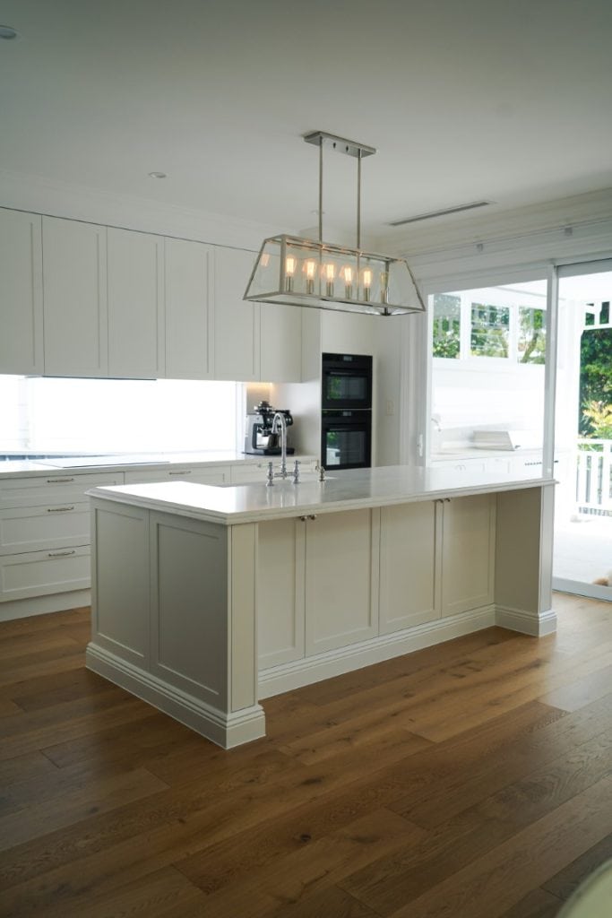 A modern kitchen with white cupboards, a large island with a sink, pendant lighting above, wooden floors, built-in ovens, and large windows letting in natural light.