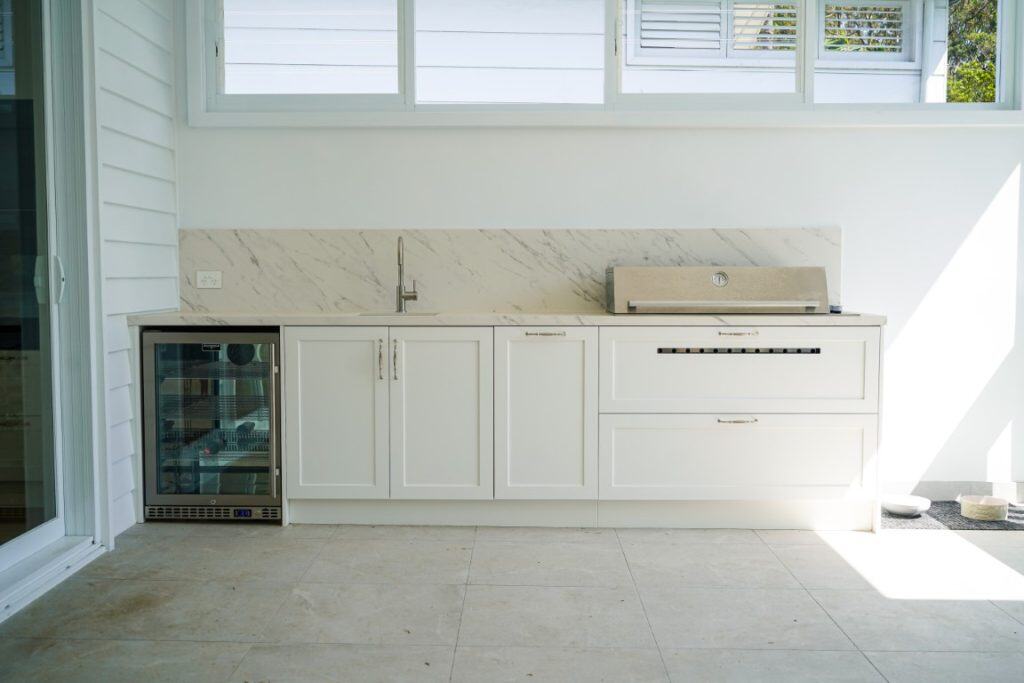 A modern outdoor kitchen with white cabinetry, a marble splashback, a built-in grill, a drinks fridge, a sink, and large windows above letting in natural light.