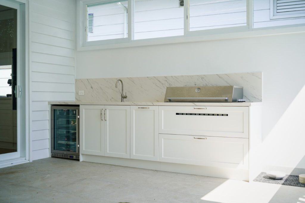 Modern outdoor kitchen with white cupboards, a marble splashback, a built-in sink, a small fridge, and a stainless steel barbecue, set against white panelled walls and under a row of windows.