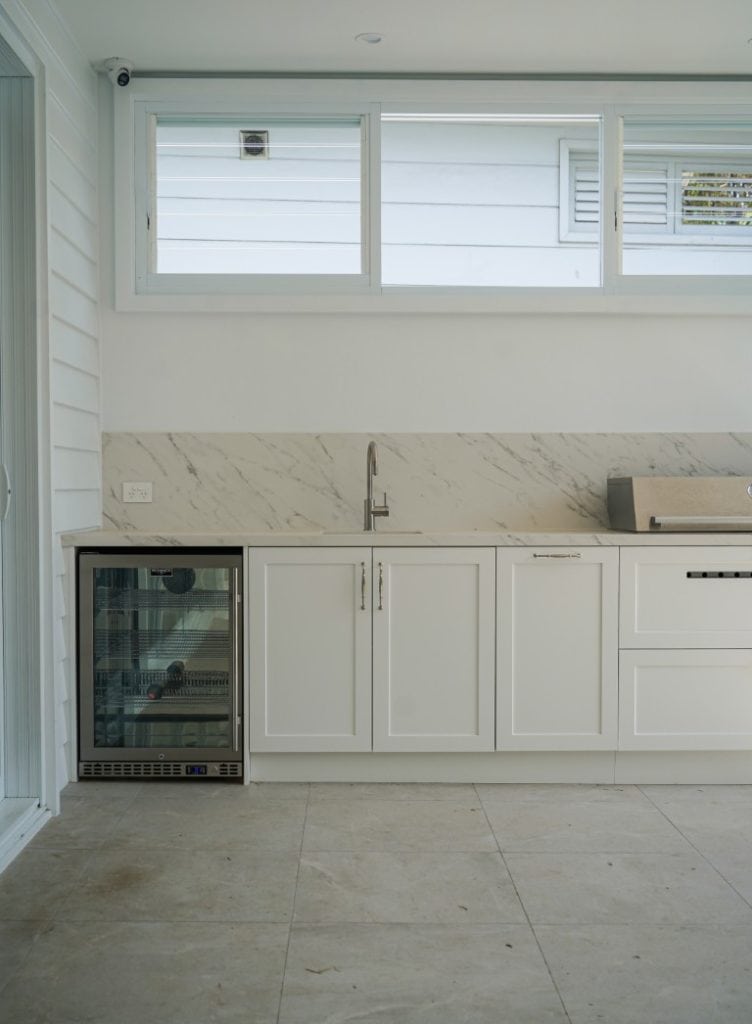 Modern kitchen with white cupboards, a marble splashback, a small sink, and a glass-fronted mini fridge under the counter. Large windows above let in natural light. The floor is tiled in light stone.