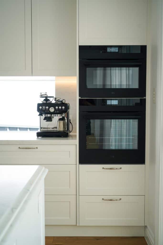 A modern kitchen corner with a built-in black oven and microwave stacked vertically, cream-coloured cabinets, and a black espresso machine on the worktop next to a window.