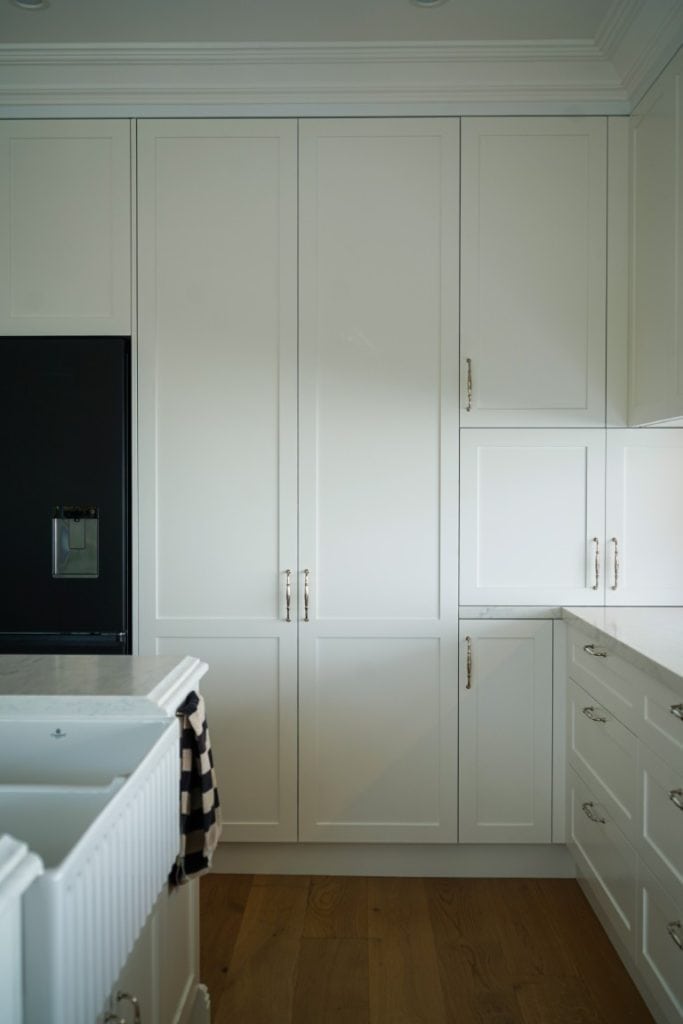 A modern kitchen with tall white cupboards, a black fridge, white worktops, and a farmhouse sink with a striped tea towel hanging over the edge. The floor is wooden, and the overall look is bright and clean.