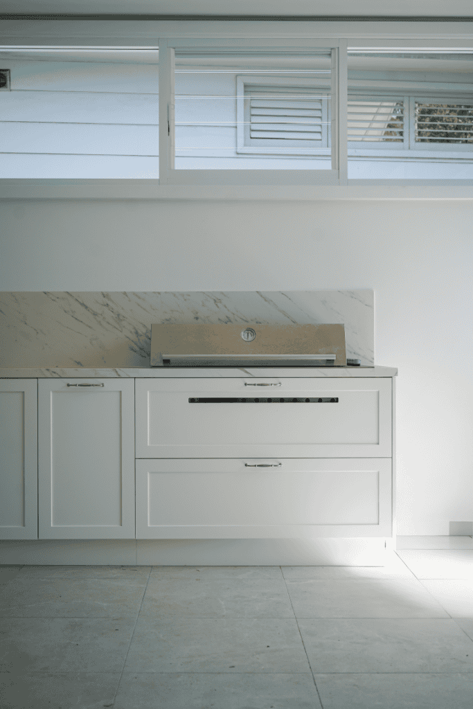 A modern, minimalist kitchen features white cabinetry, a marble splashback, and a built-in stainless steel oven, with sunlight streaming through wide horizontal windows above.