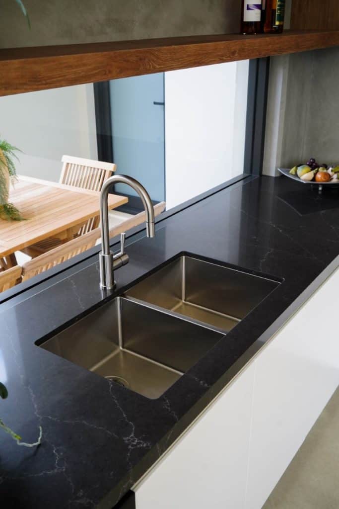 A modern kitchen with a black marble worktop, stainless steel double sink, and a sleek tap. In the background, there is a wooden dining table and a bowl of fruit on the counter.
