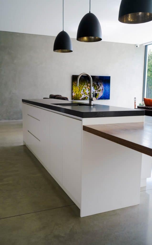 Modern kitchen with a white island featuring a black worktop and built-in sink, pendant lights above, and a minimalist design with concrete floor and walls; a TV and window are visible in the background.