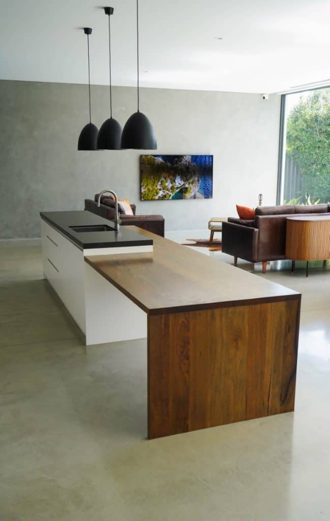 Modern kitchen with a white island, black worktop, and attached wooden dining table. Three black pendant lights hang above. In the background, a living area with sofas and a wall-mounted TV is visible.