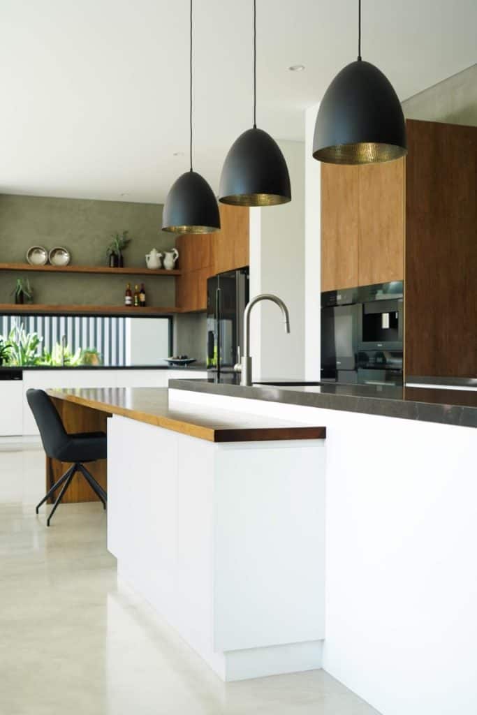 Modern kitchen with white worktops, wooden cupboards, and three large black pendant lights. There is a built-in oven, open shelves with decor, a black chair, and plants by the window adding greenery.