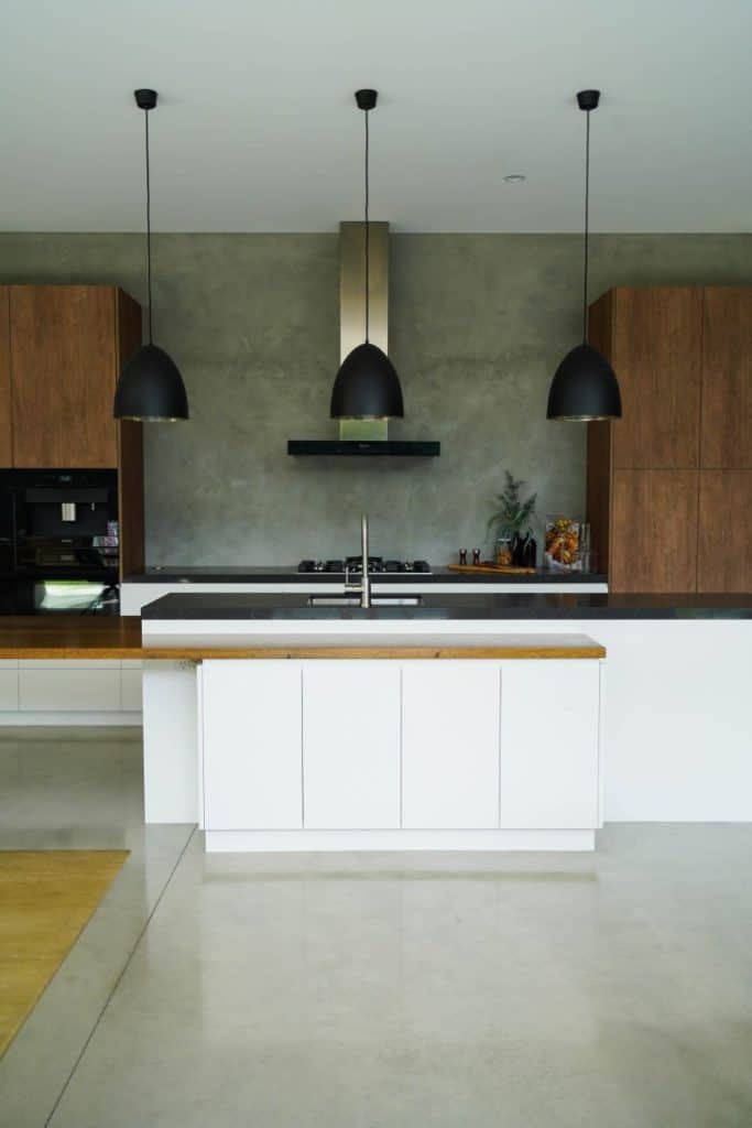 Modern kitchen with a central island, white and wood cabinetry, three black pendant lights, stainless steel appliances, and a grey concrete splashback wall. The floor is polished concrete.