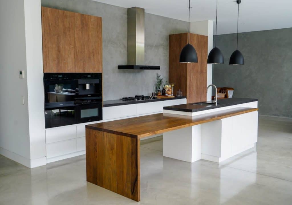 Modern kitchen with white cupboards, wood accents, a large wooden breakfast bar, stainless steel appliances, black pendant lights, and a grey concrete wall and floor. The space is bright and uncluttered.