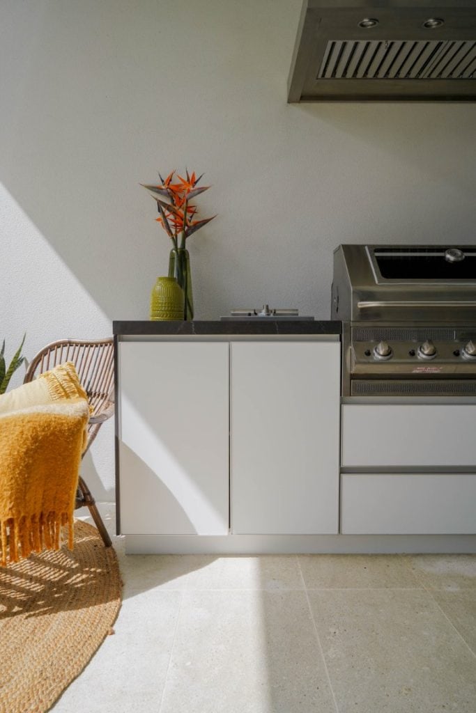 Modern outdoor kitchen with a stainless steel barbecue, white cupboards, a wicker chair with a yellow throw blanket, a round woven rug, and a vase with orange flowers on the worktop. Sunlight casts shadows across the scene.