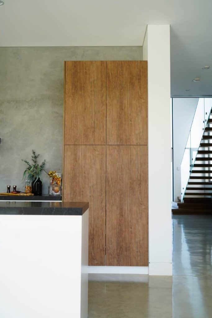 Minimalist kitchen with tall wooden cupboards, a white island with a dark worktop, concrete floor, and a modern staircase in the background; some decorative items sit atop the island and cupboard.