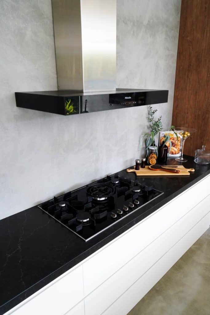 Modern kitchen with a black gas hob, black worktop, stainless steel cooker hood, and wooden accents. Cooking utensils, bottles, and a plant are neatly arranged on the counter.