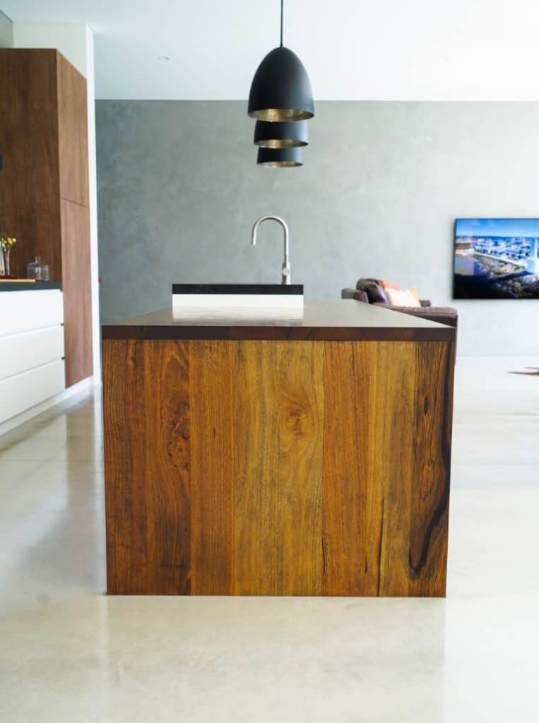 A modern kitchen features a wooden island with a sink and tap, light polished floors, pendant lights above, and a neutral grey wall in the background with a mounted TV and a sofa.