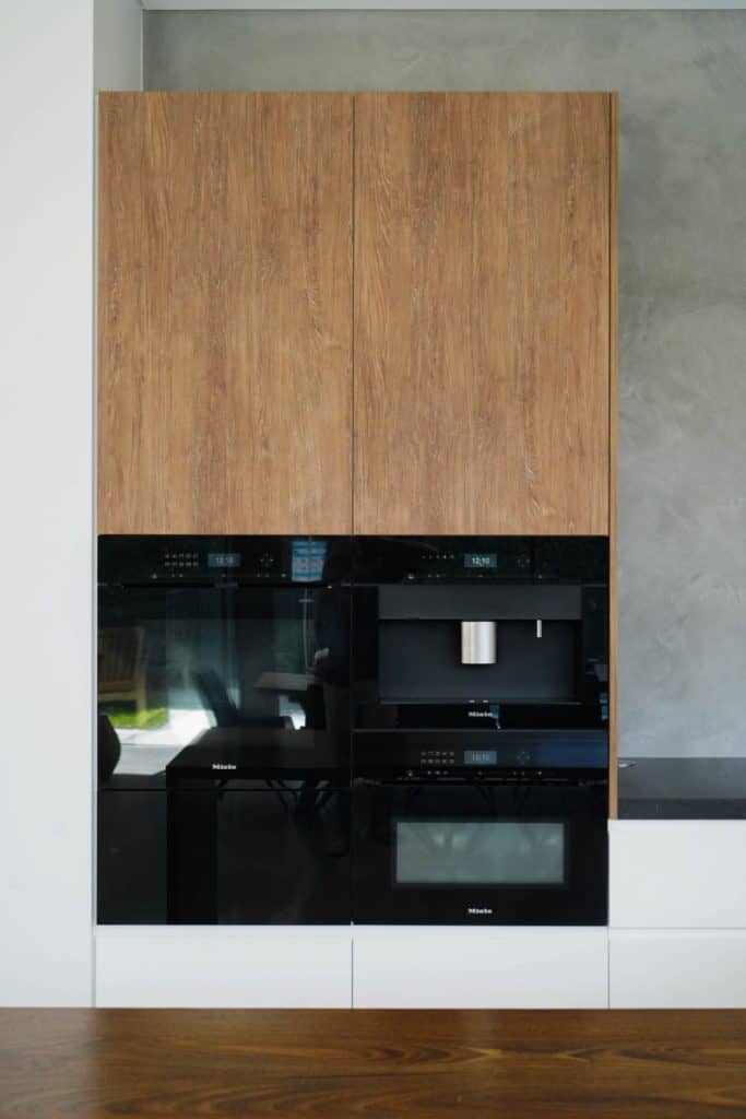 Built-in kitchen appliances with wooden cupboard doors above and sleek black glass-fronted ovens and a coffee machine below, set against a grey wall.