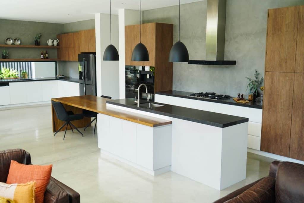 Modern kitchen with a large island, black worktops, wood and white cabinetry, hanging black pendant lights, a stainless steel cooker hood, and built-in appliances, with a black chair and plants adding decor.