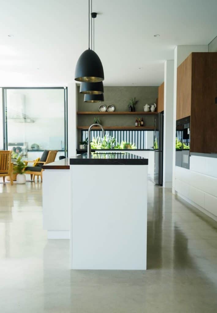Modern kitchen with a white island, black worktops, and sleek pendant lights. Wooden cupboards, open shelving, and large glass doors lead to a bright living area with plants and wooden furniture.