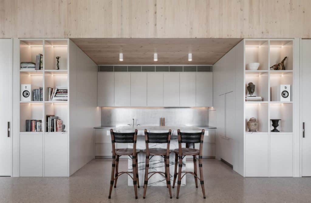 Minimalist kitchen with light wood ceiling, white cabinetry, open shelving displaying books and decor, a marble worktop island, and three wooden bar stools. Soft lighting highlights the clean, modern design.