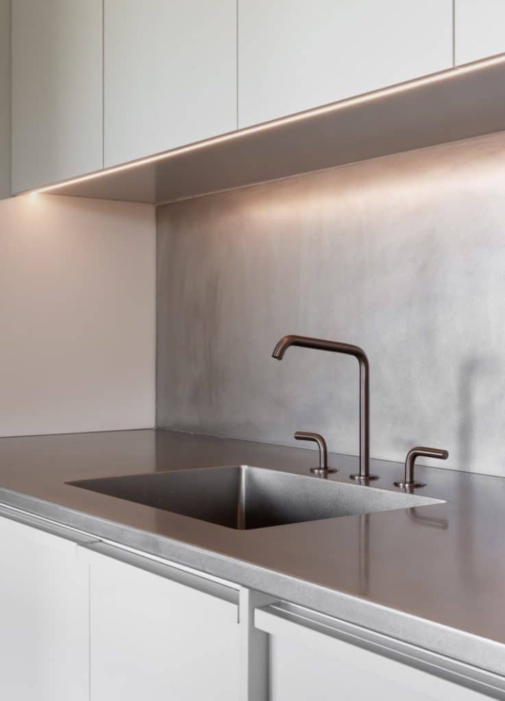 Minimalist modern kitchen with a stainless steel worktop, matching splashback, and integrated sink. Sleek, black taps are installed, and under-cabinet lighting softly illuminates the area.