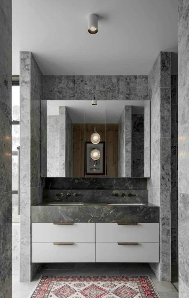 Modern bathroom with grey marble walls, a large mirror, a double sink vanity with white drawers and brass handles, and a colourful patterned rug on the floor. Two pendant lights are reflected in the mirror.