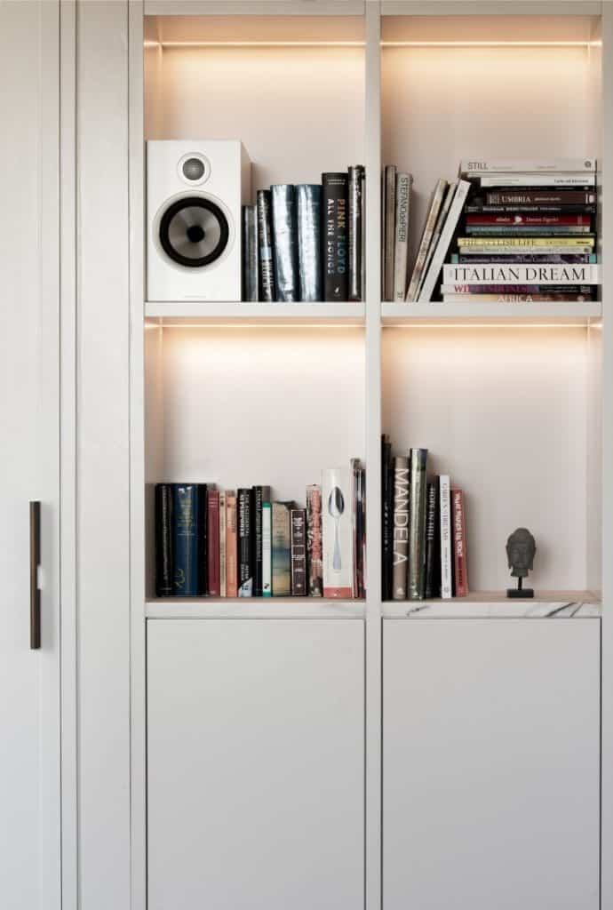 A built-in white bookcase with two shelves holding books, a white speaker, a small statuette, and some stacked magazines, all illuminated by soft, warm lighting.