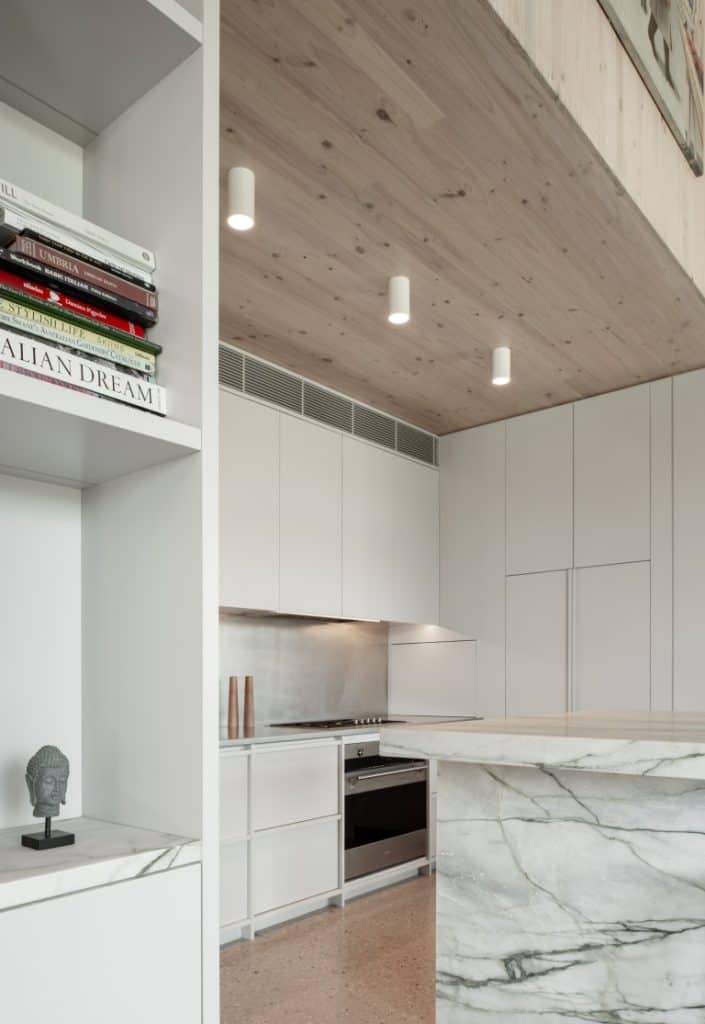 Modern kitchen with white cupboards, marble worktop and splashback, wooden ceiling with recessed lights, and a built-in shelf displaying books and a small sculpture on the left.