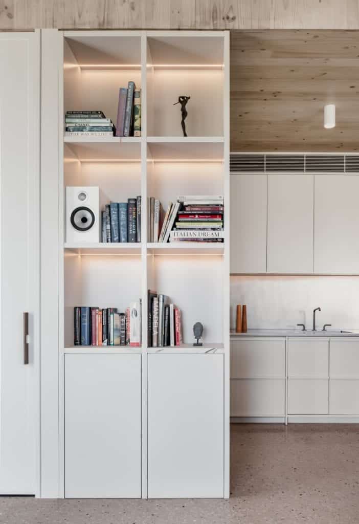 A modern white shelving unit with books, a speaker, and small sculptures, partially dividing a living space from a minimalist kitchen with light wood accents and a sink.