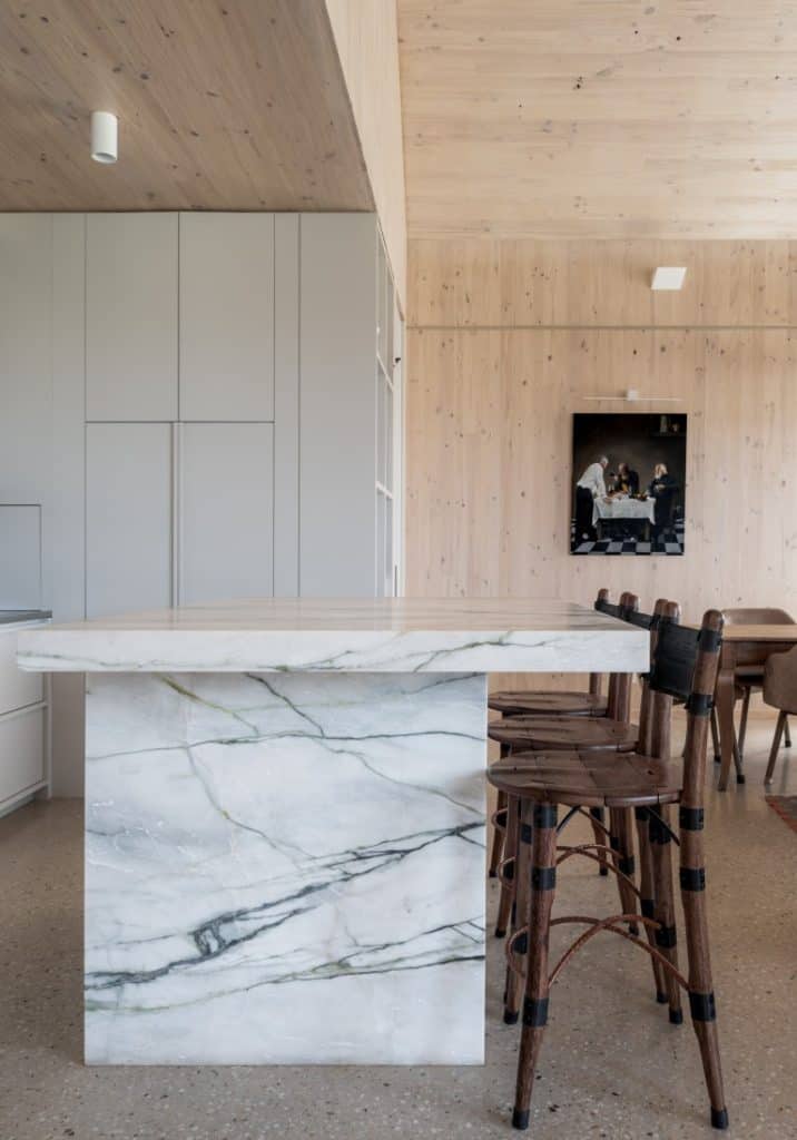 A modern kitchen with a marble island and three wooden bar stools, featuring light wood-panelled walls and a dining area with a painting in the background.