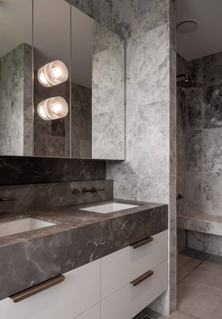Modern bathroom with grey marble walls, a large mirror, double basins with dark marble worktops, white cupboards with brown handles, and a glass-walled shower in the background. Two round lights reflect in the mirror.