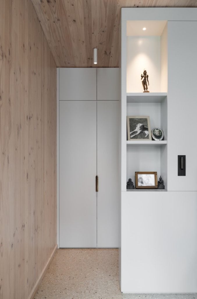 A minimal hallway with light wood walls and ceiling, terrazzo flooring, and a white built-in shelf displaying statues, framed photos, and a small clock, next to a closed white door.
