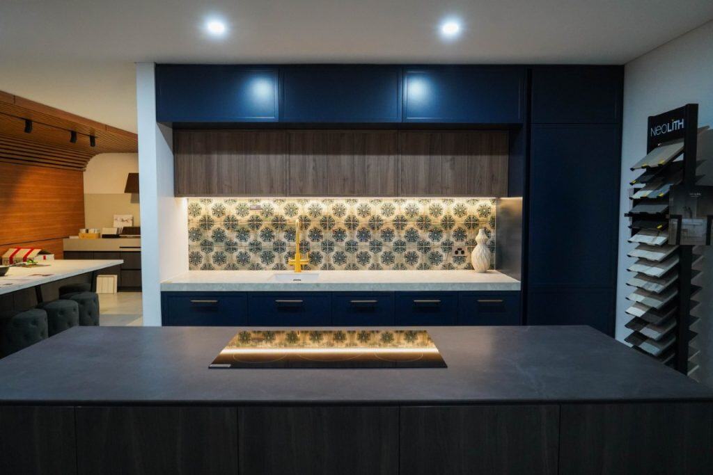 Modern kitchen with dark blue cupboards, patterned tile splashback, and under-cupboard lighting. A kitchen island with a dark worktop is in the foreground. Samples are displayed on a stand to the right.