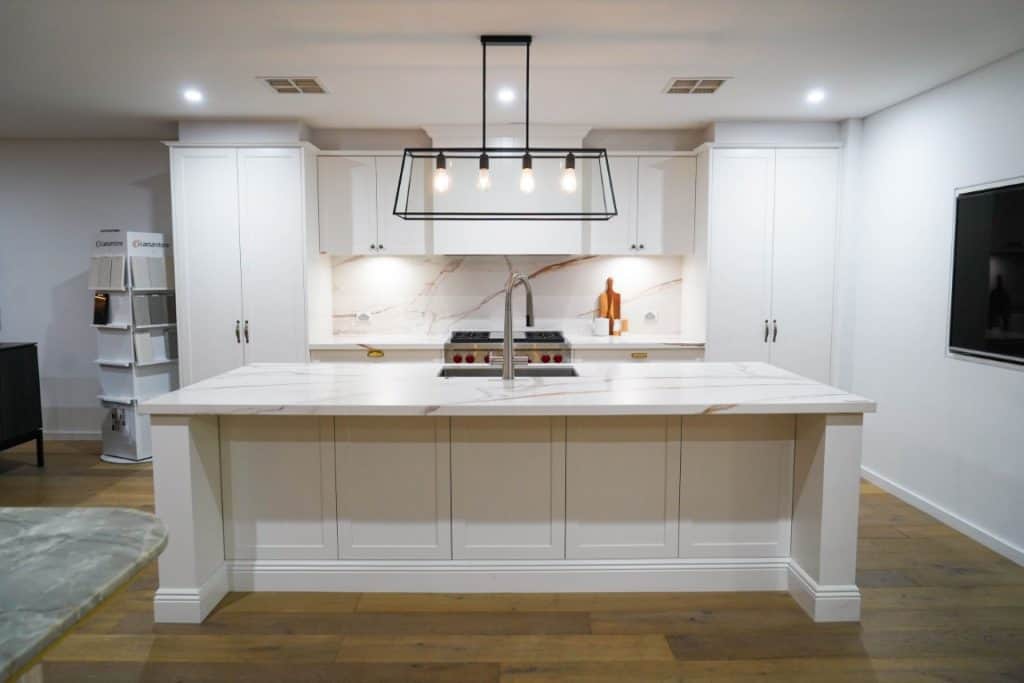 A modern kitchen with white cupboards, a marble island and worktop, a built-in cooker with an extractor hood, a sink with a tall tap, pendant lighting, and wooden flooring.