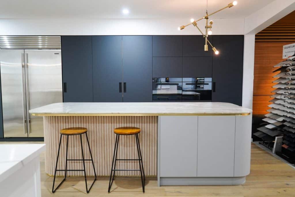 Modern kitchen with a large island featuring two wooden bar stools, sleek dark cabinetry, stainless steel fridge, built-in oven, and a contemporary gold pendant light fitting overhead.