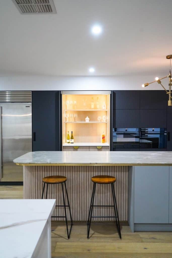 Modern kitchen with a marble island, two bar stools, dark cupboards, stainless steel fridge, and a built-in illuminated glass cabinet displaying glasses and bottles. Contemporary light fitting visible on the right.