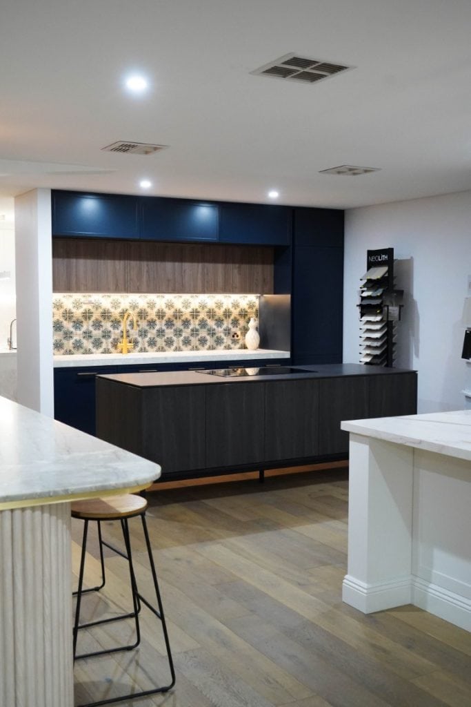 Modern kitchen with dark cupboards, a patterned tile splashback, a yellow tap, and light wood flooring. There is a white worktop island with a wooden stool and a display rack with samples against the wall.