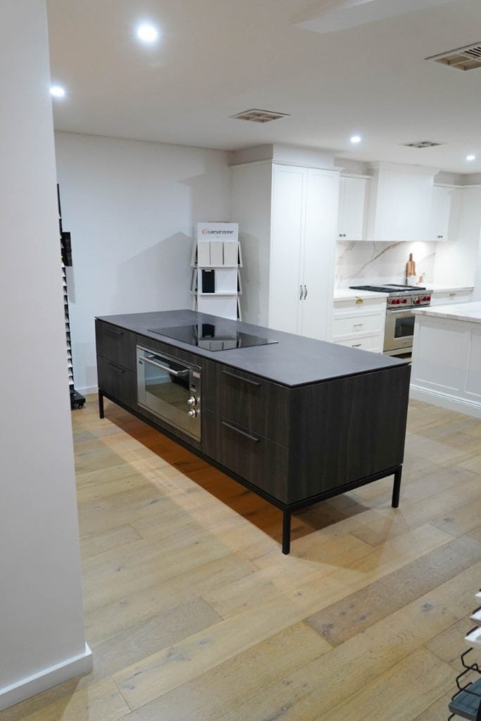 Modern kitchen with light wood flooring, a dark island featuring an electric hob and built-in oven, and white cupboards in the background. The space is well-lit and minimalist in design.