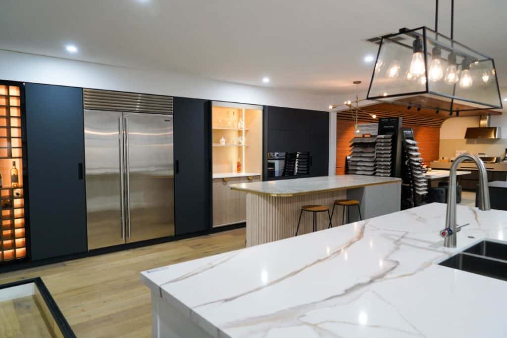 Modern kitchen with marble worktops, a large island with two bar stools, stainless steel double-door fridge, dark cabinetry, open shelves, and pendant lighting. Warm wooden flooring and ambient lighting create a cosy atmosphere.