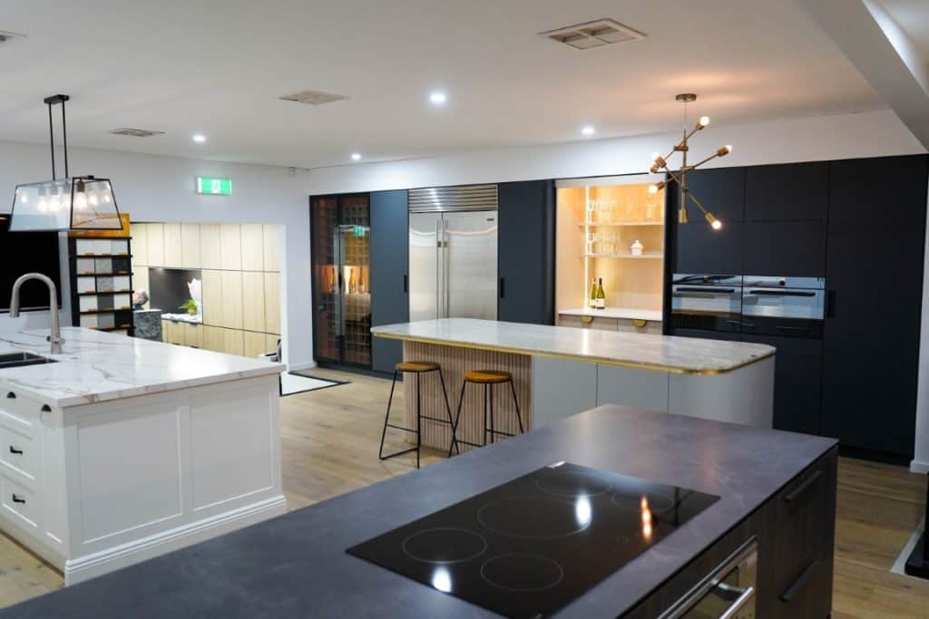 Modern kitchen with marble worktops, a large island with two stools, sleek appliances, built-in shelves, pendant and modern ceiling lights, and wooden flooring. An open area with a green exit sign is visible in the background.