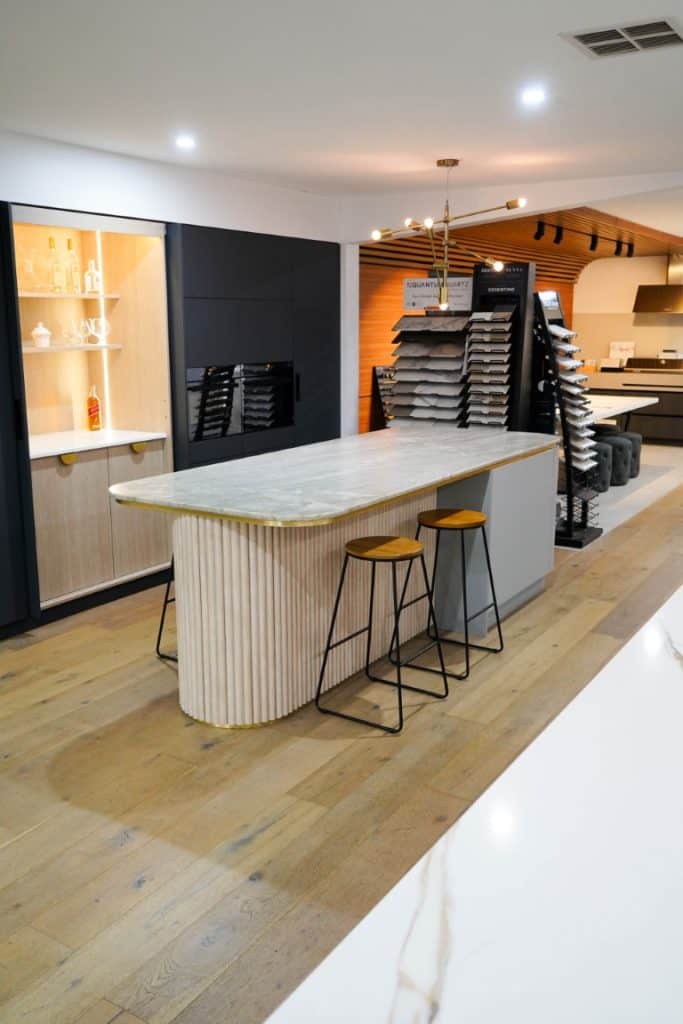 Modern kitchen showroom with a marble island, two bar stools, wood flooring, display shelves with glassware, and sample boards stacked on a side counter. Warm lighting highlights the contemporary design.