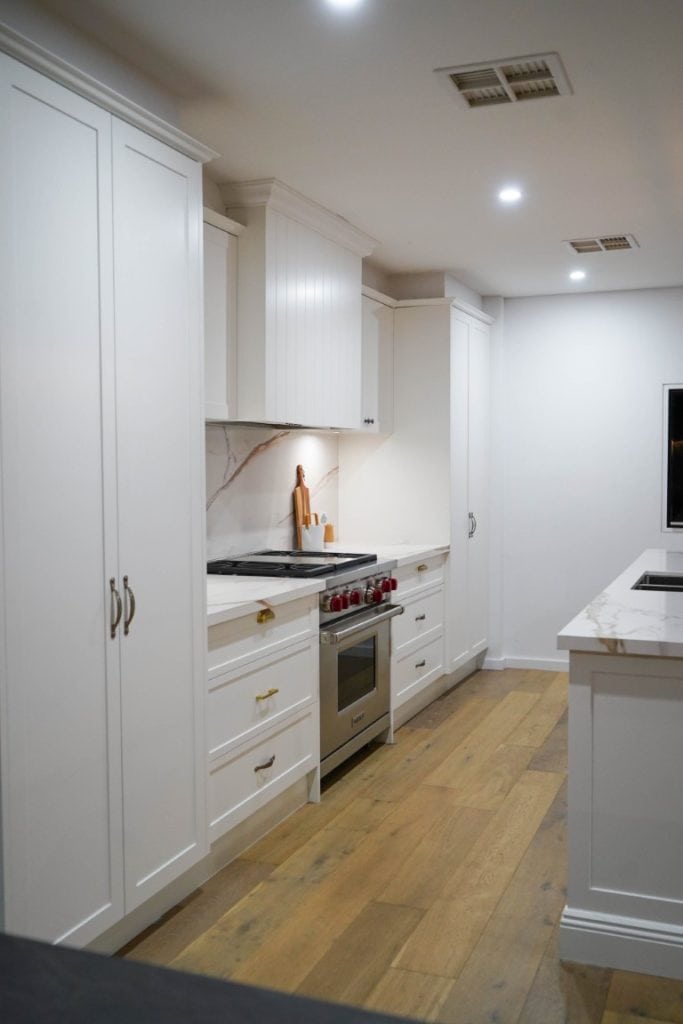 Modern kitchen with white cupboards, stainless steel oven, light wood flooring, marble splashback and worktops, and recessed ceiling lights. A chopping board and utensils are on the worktop by the cooker.