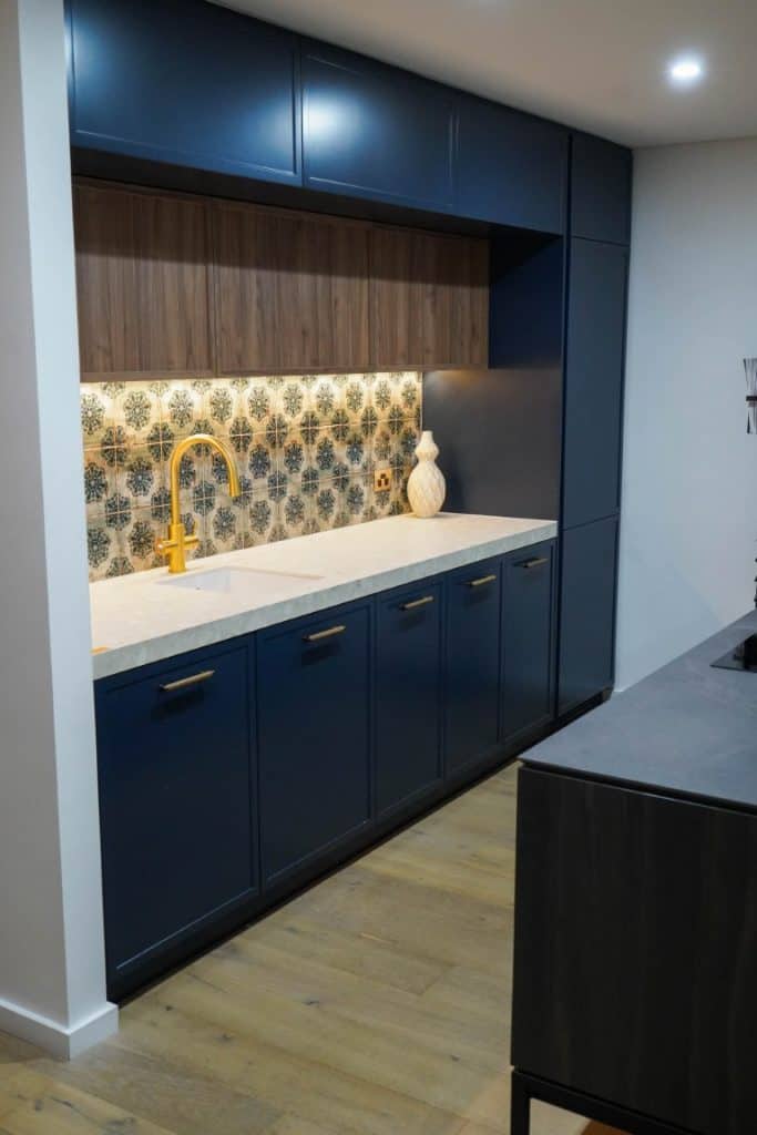 A modern kitchen with dark blue cupboards, a white worktop, a gold tap, patterned tile splashback, wood accents, and decorative vases on the counter. Warm under-cupboard lighting highlights the splashback.
