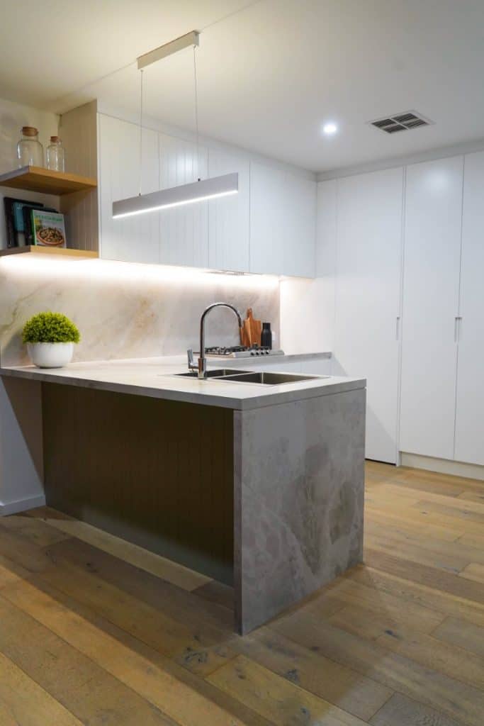 Modern kitchen with light wood floors, white cupboards, a marble worktop and splashback, built-in sink, potted plant, cookery books on an open shelf, and minimalist hanging light fitting.