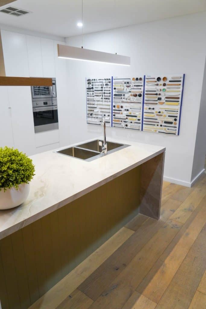 Modern kitchen with a marble island worktop, built-in stainless steel sink, potted plant, wooden flooring, and white cupboards. Display boards with various cupboard handles and knobs are mounted on the wall.