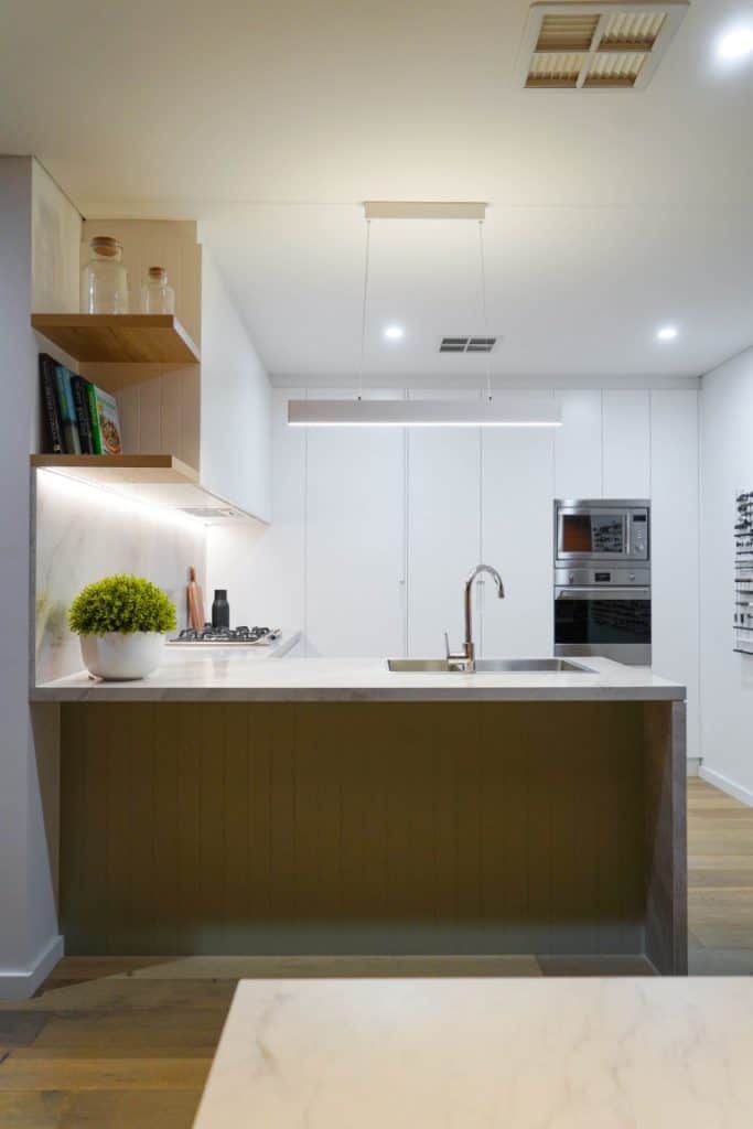 Modern kitchen with a marble worktop island, built-in sink, hob, oven, and microwave. Open shelves with jars and plants, under-cabinet lighting, and minimalistic white cupboards are visible.