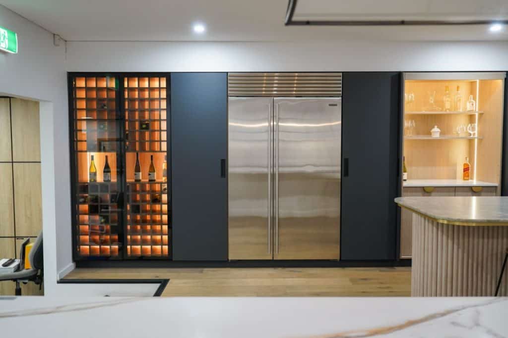 Modern kitchen with a large stainless steel double-door fridge in the centre, a backlit wine rack on the left, and a glass display cabinet with shelves and drinks on the right. Marble worktop in the foreground.