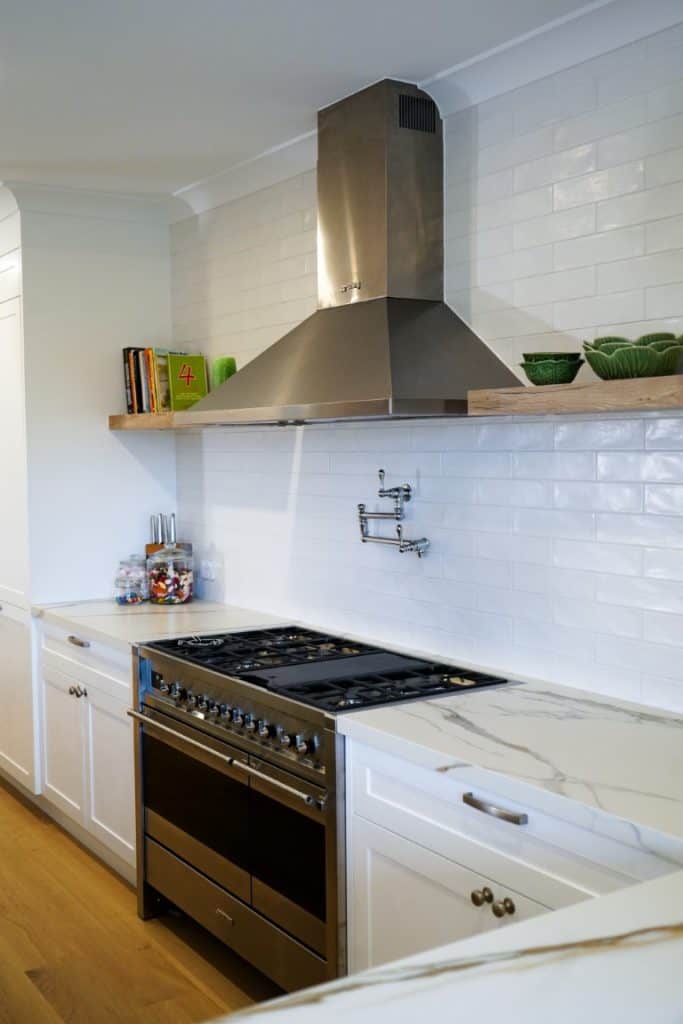 Modern kitchen with white cupboards, marble worktops, a stainless steel gas cooker and extractor hood, a wall-mounted pot filler, floating shelf with green bowls, and cookery books against a white metro tile splashback.