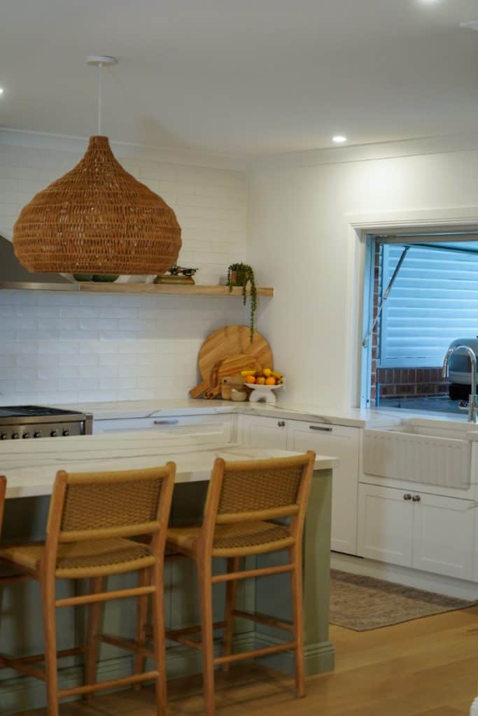 A modern kitchen with white cupboards, a farmhouse sink, a marble worktop, two wicker bar stools, a woven pendant light, open shelving with decor, and a window above the sink.