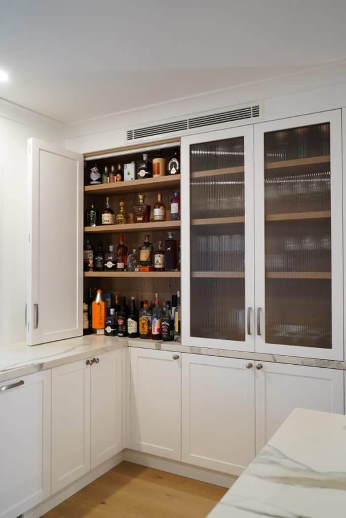 A kitchen corner cupboard with white doors, one open to reveal shelves stocked with various bottles of liquor and spirits. Adjacent cupboards have textured glass doors. White worktops and wooden floor are visible.