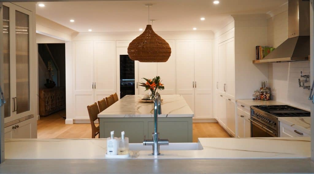 A modern kitchen with white cupboards, a large island with seating, a wicker pendant light, stainless steel appliances, and a vase with flowers on the island. The space is brightly lit with recessed lighting.