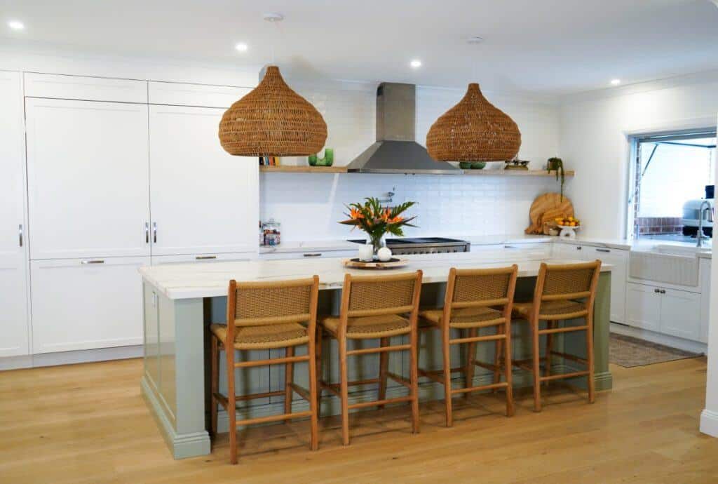 Modern kitchen with a large island, four woven bar stools, two wicker pendant lights, white cupboards, stainless steel appliances, a vase of flowers on the island, and wooden flooring.